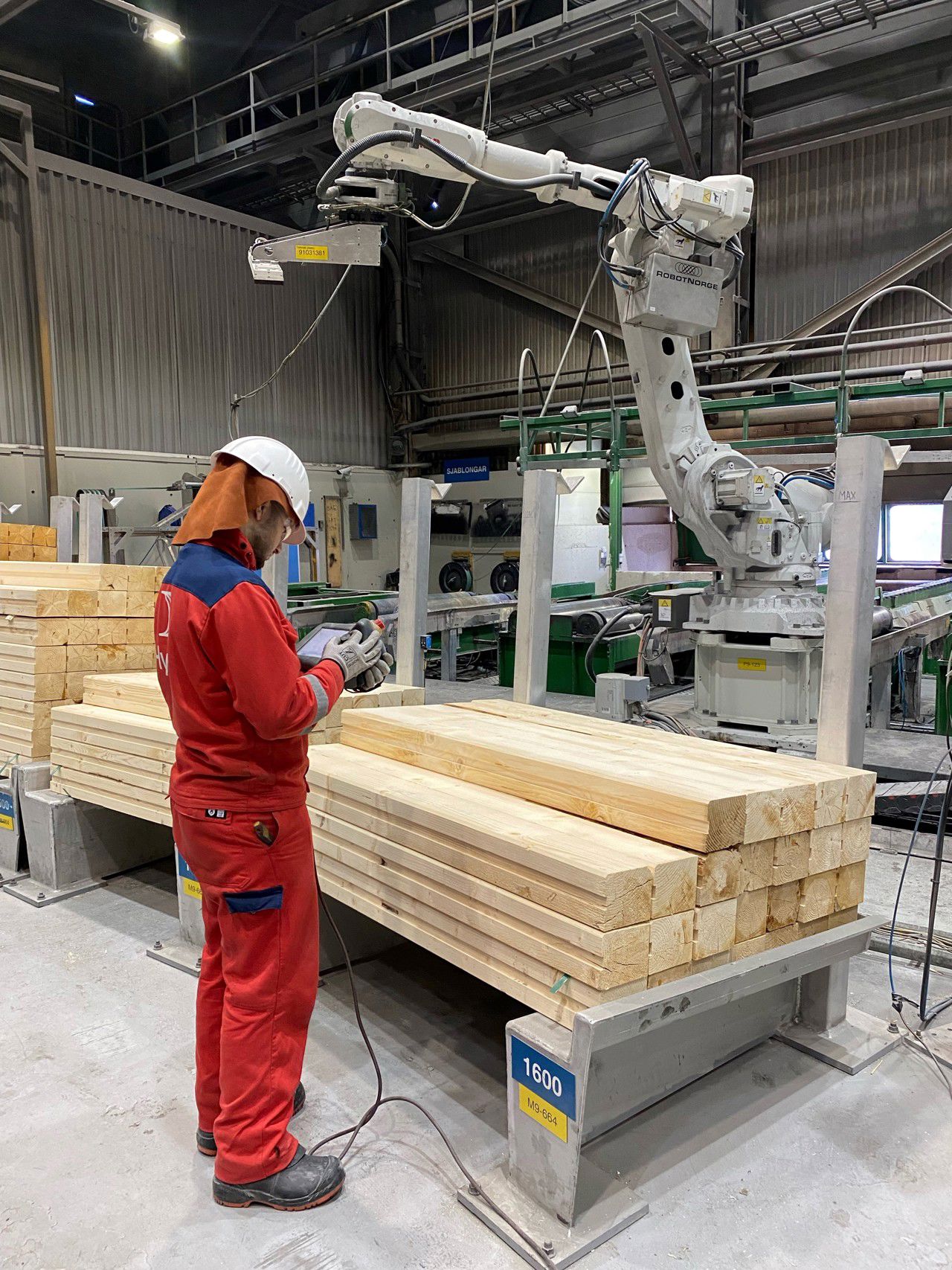Elev frå vg3 automatisering på utplassering i Støyperi Tya på Hydro Aluminium i Årdal. Han står iført arbeidsklede og verneutstyr. Han står i ein stor hall med eit kontrollpanel i handa. Rundt han er det ein stablar med trelast og ein stor robot.