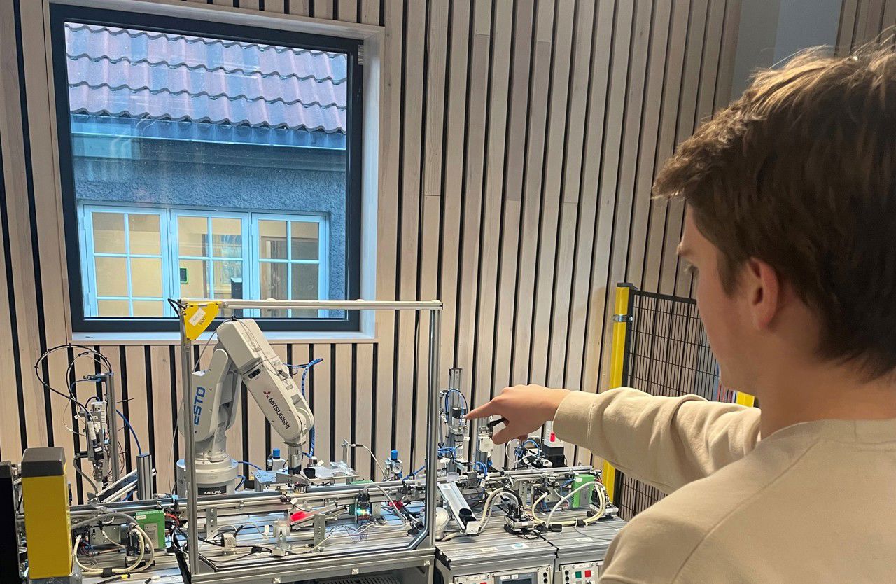 Ein elev frå automatiseringsfaget demonstrerer kva han har koda og programmert den eine roboten i Årdal teknologipark til å utføre. I bakgrunnen er det spilevegg med eit vindauge der ein kan sjå bygget på andre sida. I fokus peiker elevn mot roboten som utfører instruksjonane sine. 