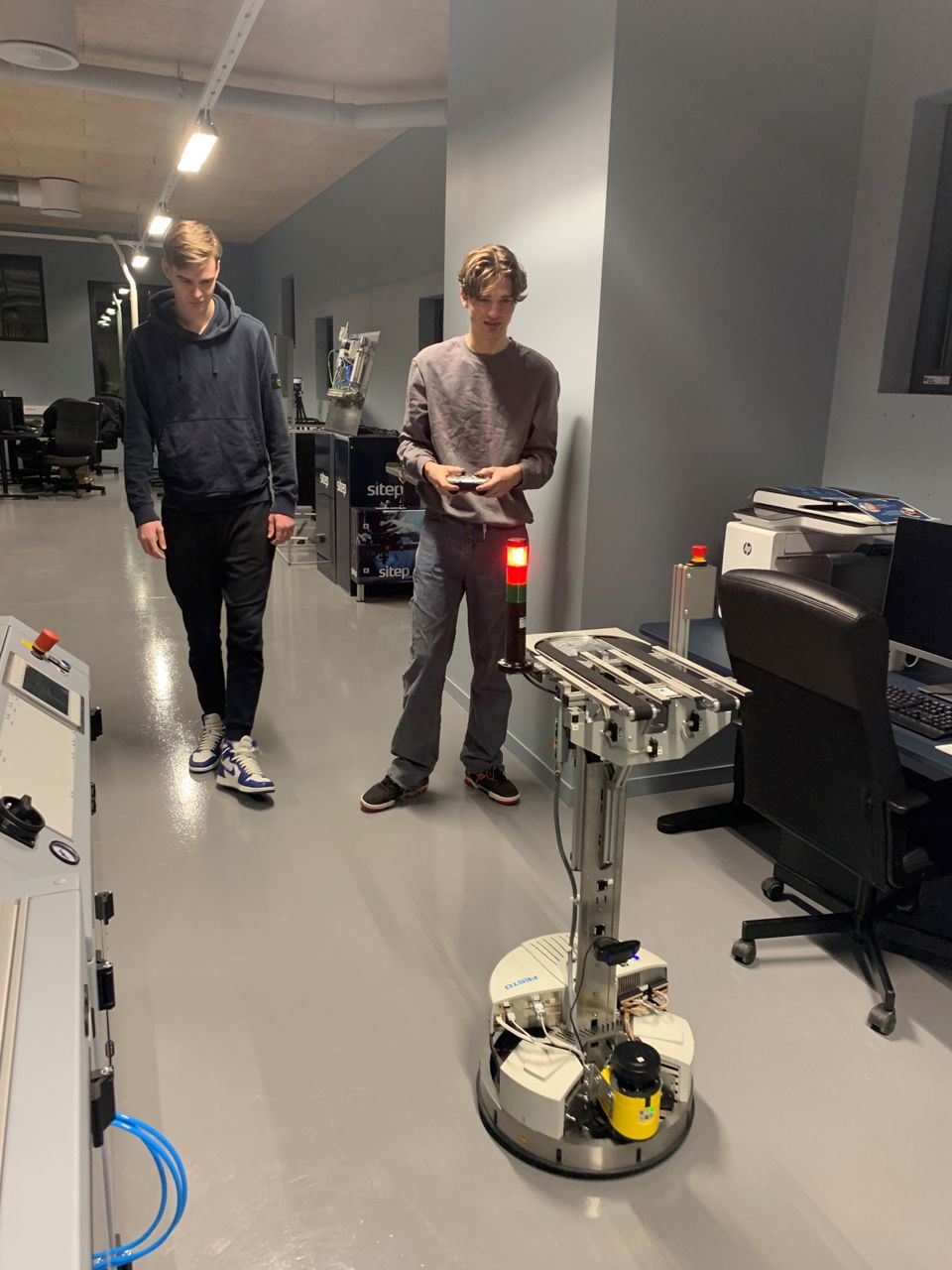 To elevar frå automatiseringsfaget styrer ein av robotane i Årdal teknologipark. Ein av dei held ein fjernkontroll i handa, medan den andre står ved sidan av. Roboten er i fokus. 