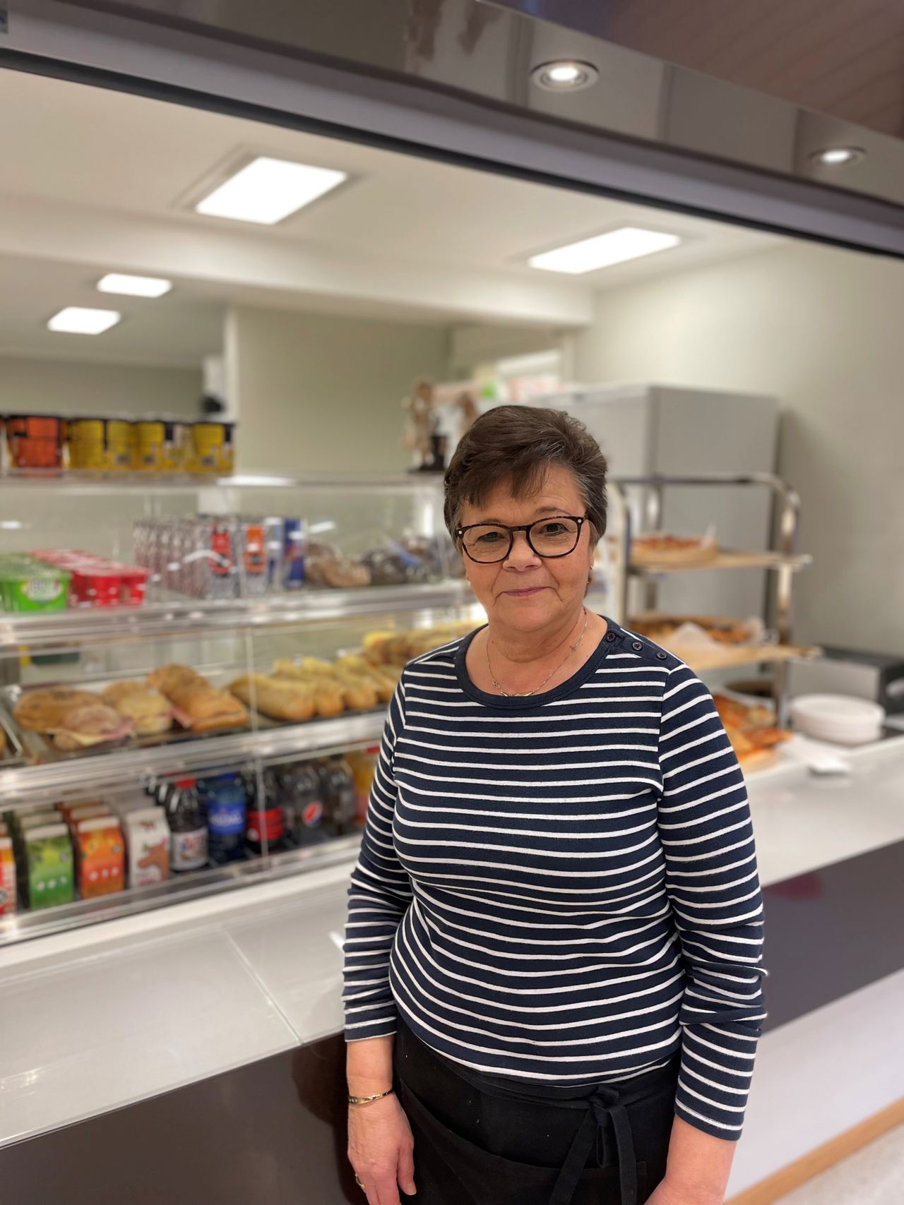 Margot som driftar kantina på Årdal vgs står framfor disken. I bakgrunnen er det kjøledisk med drikke, yoghurtar og diverse påsmurte baguettar. 