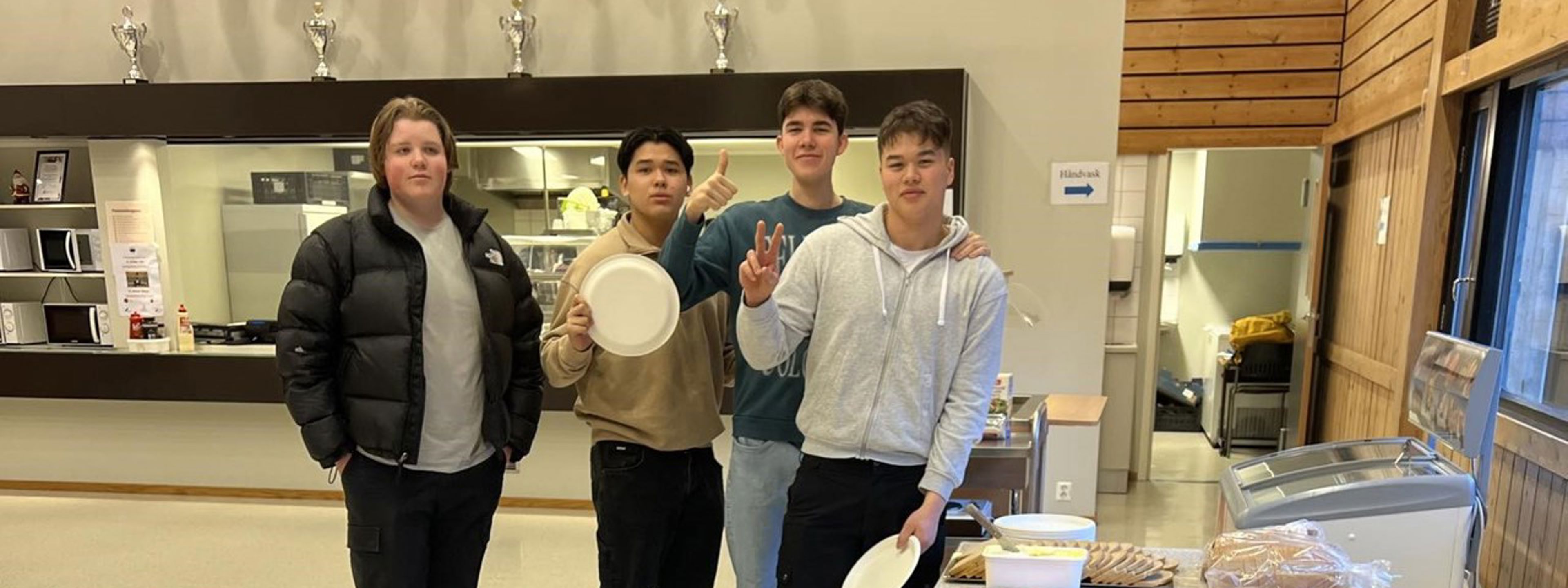 Fire gutar som er elevar ved skulen smiler fint til fotografen i kantina. Dei står ved sidan av langbordet der det er sett fram frukost til elevane. 