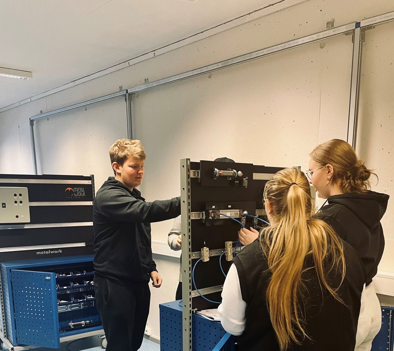 Tre elevar frå teknologi- og industrifag som koblar på ein vegg. Bilete viser ryggen på to jenter og andletet til ein gut. I bakgrunnen ser du resten av arbeidsrommet.