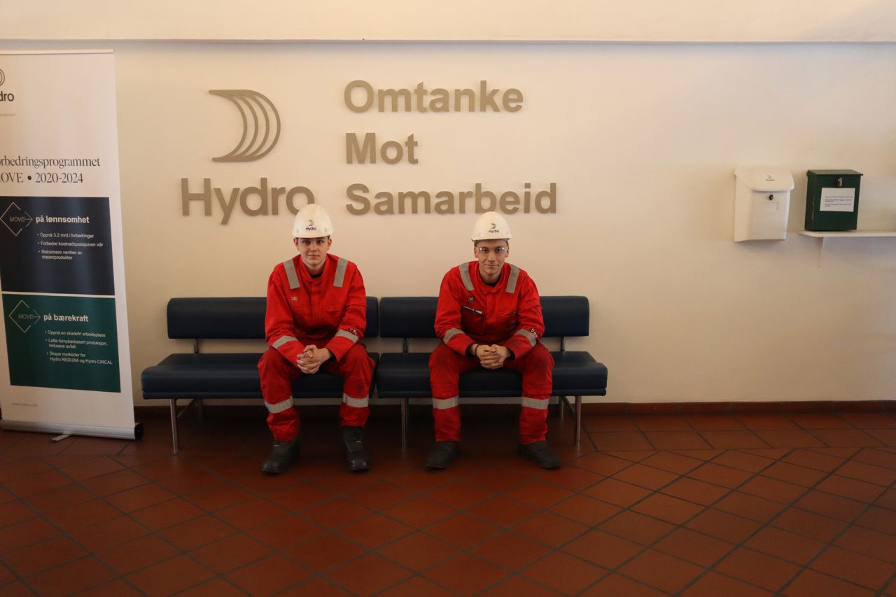  Elevar frå kjemiprosess- og laboratoriefag på utplassering hjå Hydro Årdal. Dei sit saman på ein benk. I bakgrunnen ser du logoen til Hydro med selskapet sine verdiar omtanke, mot og samarbeid ved sidan av. 