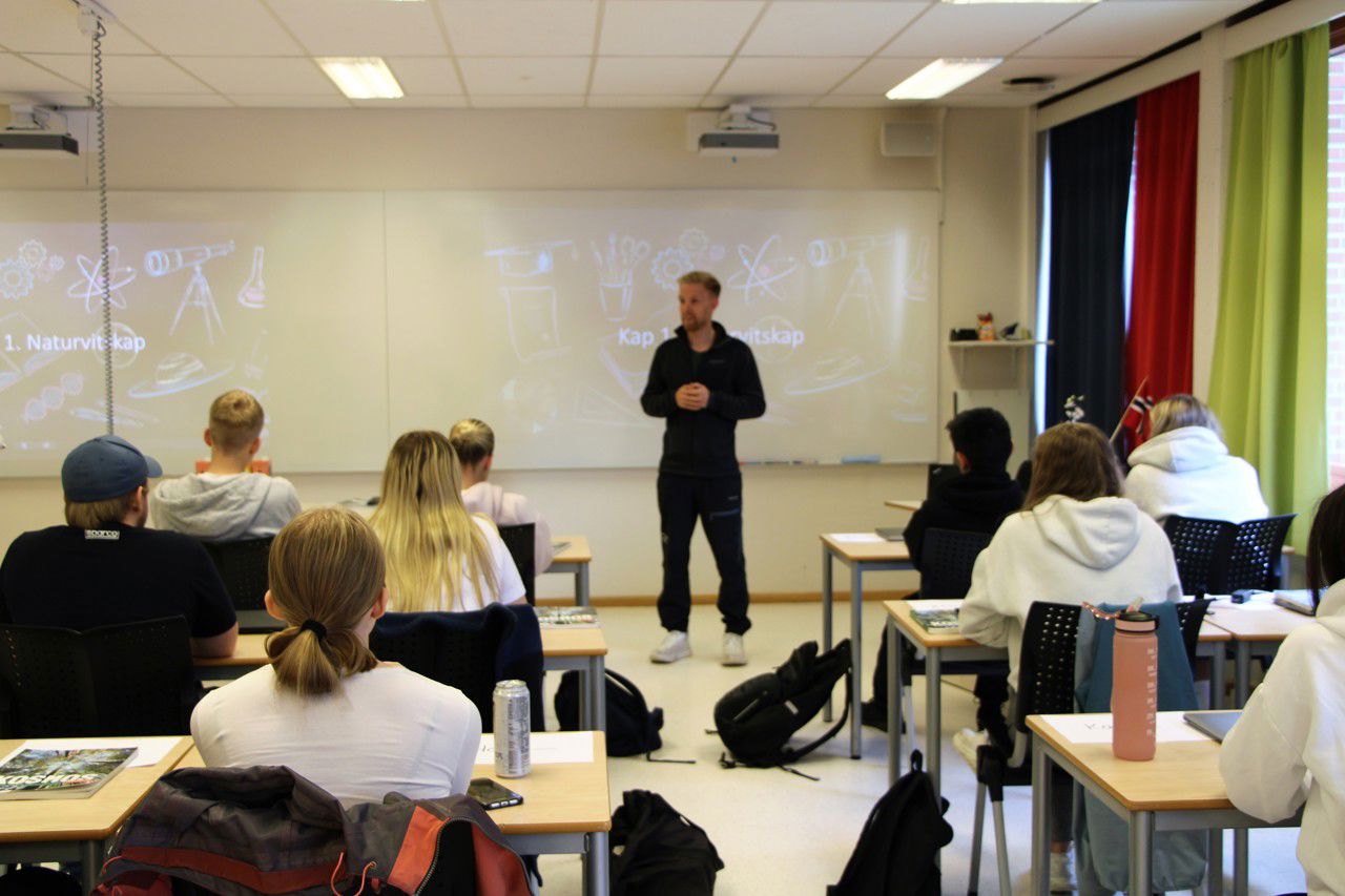 Bilete teke bakfrå i klasserommet. Du ser ryggen til elevane og ein lærar som står framme ved tavla og underviser. På tavla står det naturvitskap.