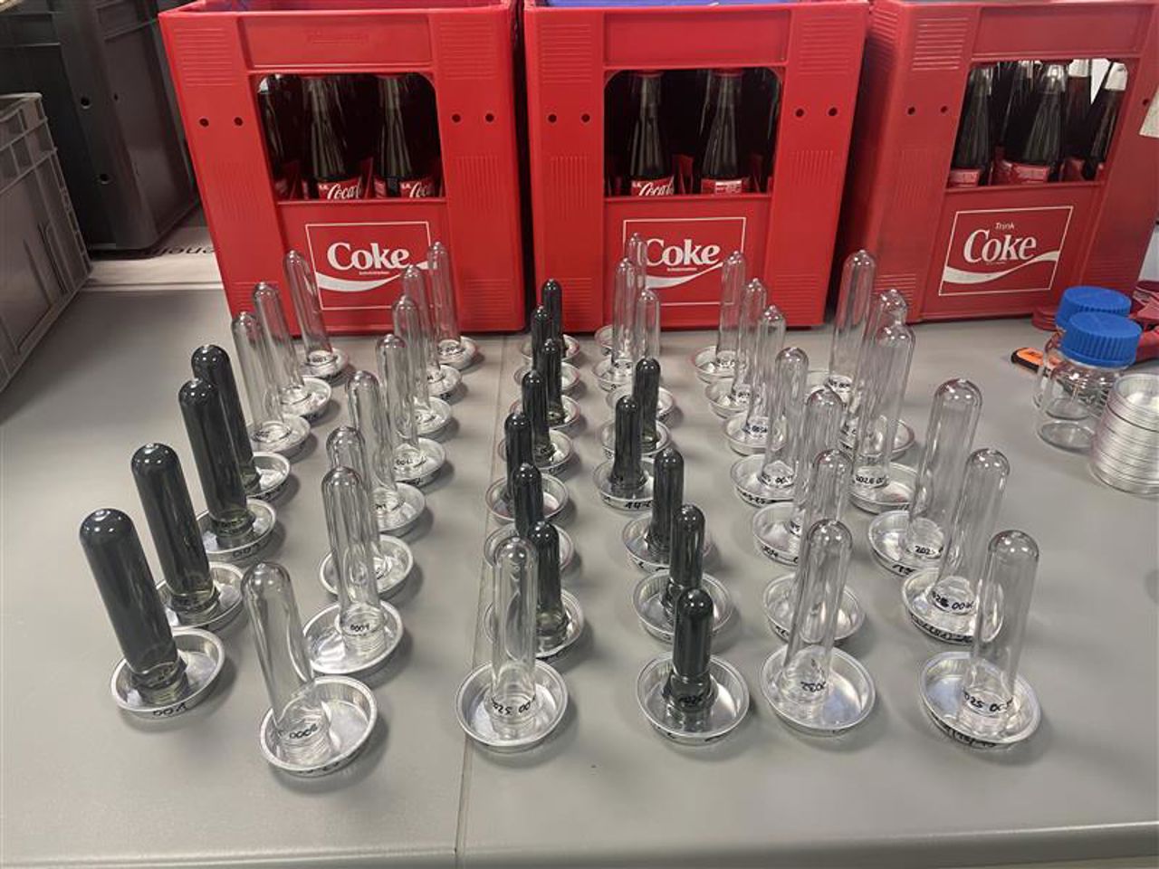 Mange reagensglas med prøvar i. Kassar med coca-cola i bakgrunn.